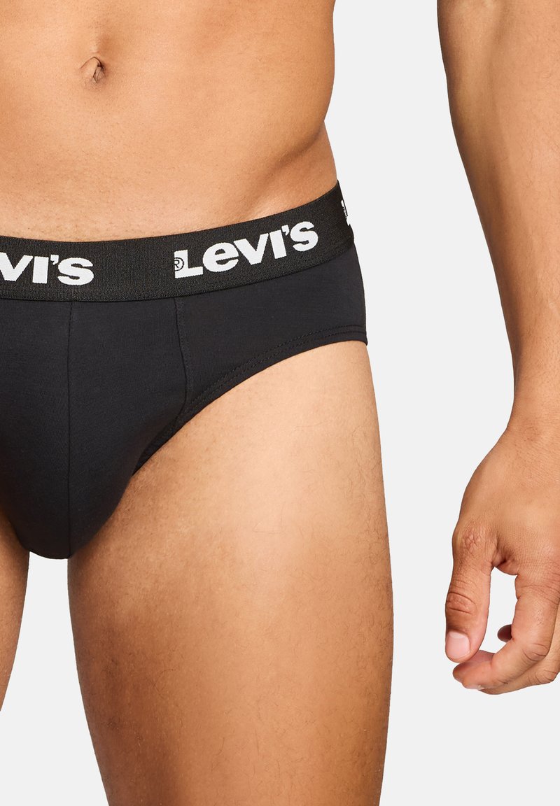 Männlicher Oberkörper und obere Beine in schwarzen Levi's-Slips mit gebrandetem Bündchen, rechte Hand entspannt an der Seite, vor einem einfarbigen Hintergrund.