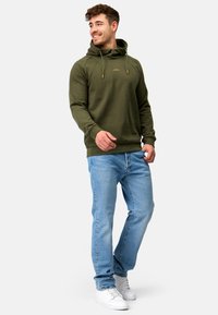 Sudadera con capucha de color verde oliva con cordones y detalles dorados, combinada con vaqueros de color azul claro y zapatillas blancas. Textura de tela suave.