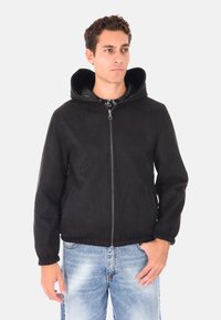 Uomo che indossa una giacca a zip nera con cappuccio e jeans chiari sbiaditi, in piedi con una mano in tasca contro uno sfondo semplice.