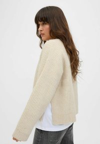 Suéter de punto acanalado beige con un ajuste relajado, hombros caídos y aberturas laterales. Llevado sobre una camiseta blanca, combinado con jeans grises.