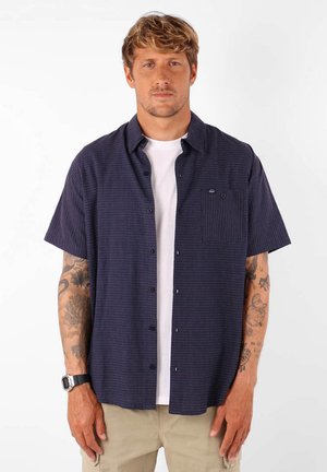 Chemise à manches courtes et boutonnée en marine avec un motif texturé, poche poitrine et un t-shirt blanc en dessous. Le tissu semble léger et respirant.