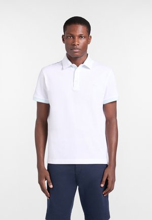Jeune homme debout immobile portant un polo blanc à manches courtes et un short bleu foncé sur un fond blanc uni.