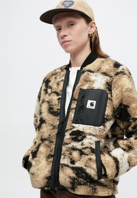 Chaqueta bomber de forro polar con un estampado de leopardo en beige y negro, que presenta un cierre de cremallera en el frente, acentos en negro y un bolsillo con parche.