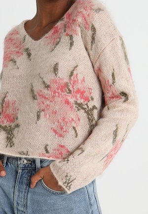 Jasnobeżowy dzianinowy sweter z różowym i zielonym kwiatowym wzorem, noszony z niebieskimi dżinsami, swobodnie ręka w kieszeni.