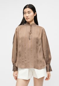 Γυναίκα με καφέ ριγέ μακρυμάνικο μπλουζάκι με στιλ γιακά και λευκά σορτς, στέκεται μπροστά σε απλό φωτεινό φόντο.