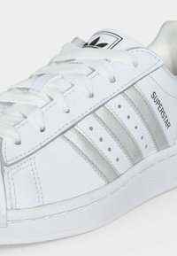 Άσπρο αθλητικό παπούτσι Adidas Superstar με ασημένιες ρίγες, λευκά κορδόνια, μαύρο λογότυπο στη γλώσσα και κείμενο "Superstar" στην πλευρά.