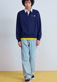 Marinblå sweatshirt med vit krage, med en färgglad 'CHIP'-logotyp; kombinerad med ljusblå jeans och svarta sneakers.
