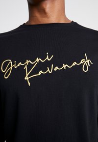 Černé tričko s námořnickou výšivkou zlatým kurzivním písmem "Gianni Kavanagh." Materiál vypadá měkce, s klasickým kulatým výstřihem a volným střihem.