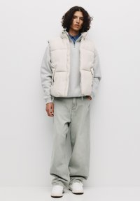 Padded beige vest over a gray sweater, paired with loose light gray jeans and white sneakers. Model stands against a plain background.