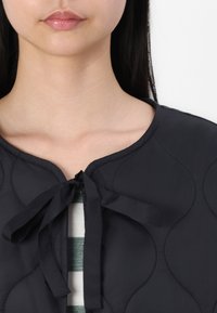 Jachetă neagră matlasată cu un guler rotund, dotată cu un sistem de închidere prin legare și un model texturat. O bluză cu dungi verzi și albe este vizibilă dedesubt.