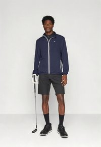 Casaco azul-marinho com fecho éclair branco e detalhes brancos, calções pretos e sapatos pretos. Modelo segura um taco de golfe, evidenciando um design desportivo.