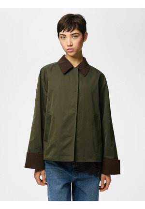 Młoda osoba ubrana w oliwkowo-zieloną kurtkę z ciemnobrązowym kołnierzem i mankietami, w połączeniu z niebieskimi dżinsami, stojąca na białym tle.