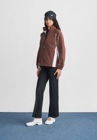 Καφέ αδιάβροχο με λευκά πλαϊνά πάνελ, φερμουάρ και ελαστικά μανσέτες. Συνδυασμένο με μαύρο παντελόνι και λευκά αθλητικά παπούτσια, ολοκληρωμένο με μαύρο καπέλο.