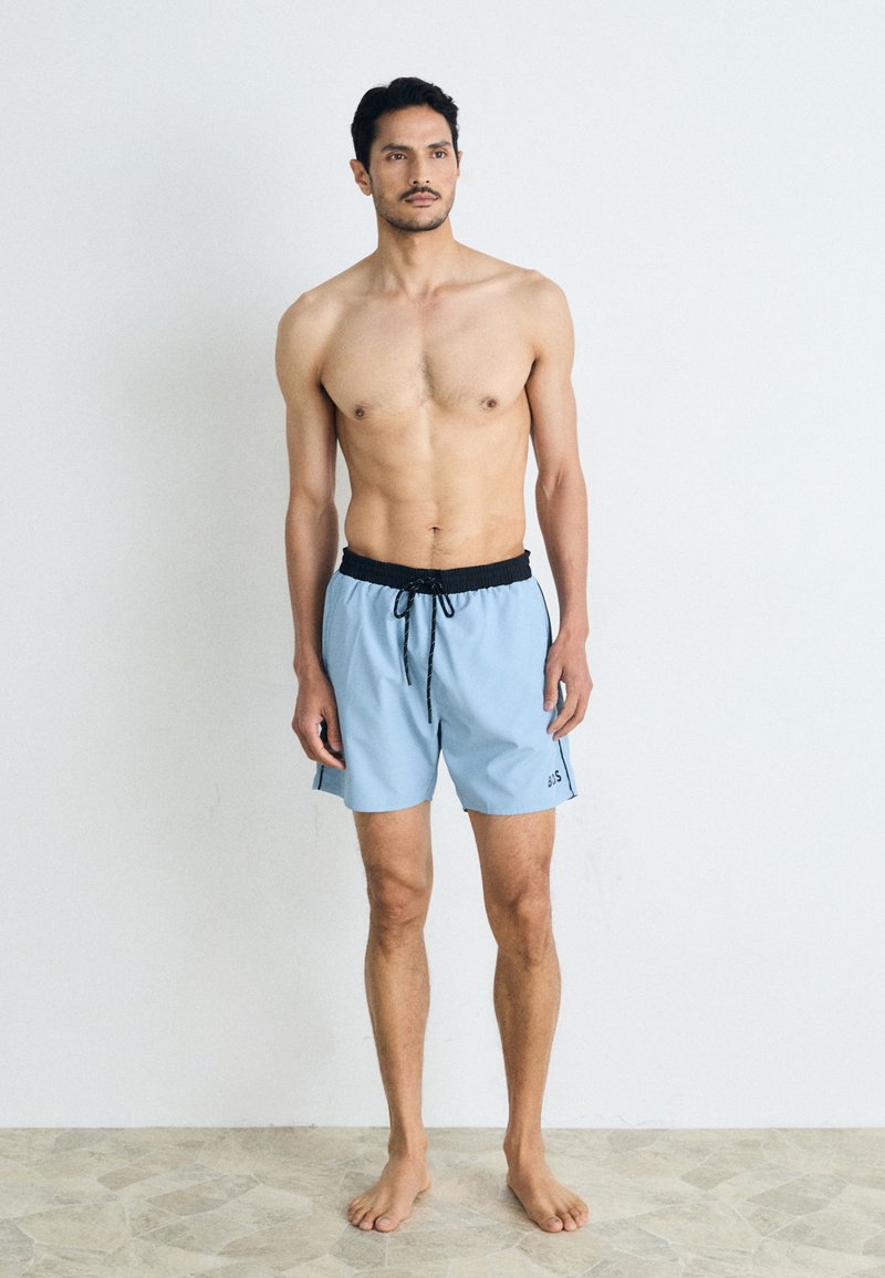 Shorts de bain bleu clair avec une ceinture élastique noire et un cordon de serrage. Présente des poches latérales et un petit logo sur la jambe gauche.