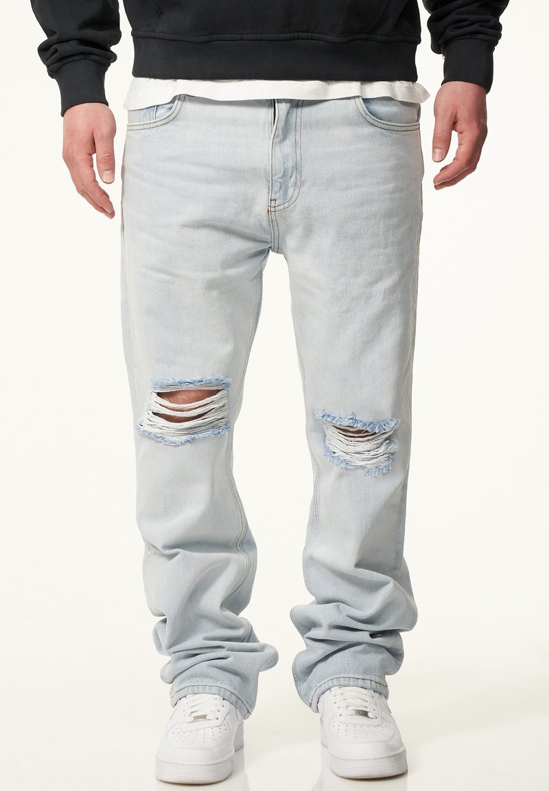 Light blue denim jeans with distressed rips at the knees, loose fit, and cuffed ankles, paired with white sneakers.