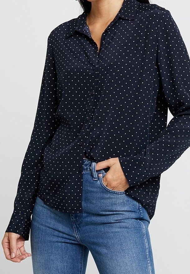 Femme portant une chemise bleu marine à manches longues avec de petits pois blancs et un jean bleu, main dans la poche devant un fond uni.