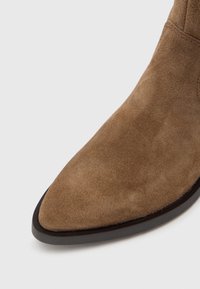 Brązowy zamszowy botek na obcasie z czarną podeszwą na jednolitym białym tle.