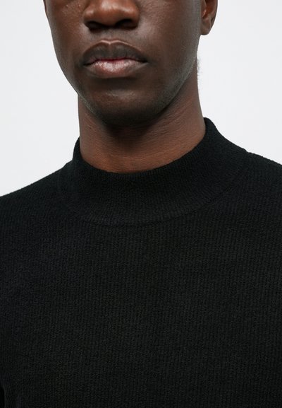 Primo piano di una persona che indossa un maglione nero a collo alto con texture, mostrando la parte inferiore del viso, le labbra e il collo su uno sfondo neutro.