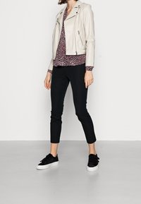 Femme portant une veste en cuir crème, un chemisier bordeaux à motifs, un pantalon slim noir et des baskets noires, se tenant contre un mur blanc.