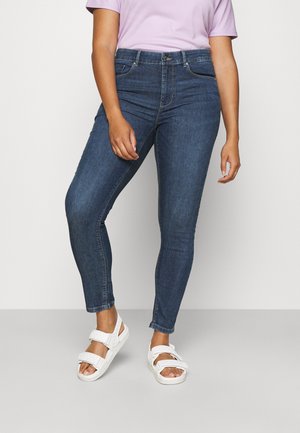 Dark blue skinny jeans made of denim, featuring a mid-rise waist, front pockets, and a subtle faded texture. Paired with white sandals.