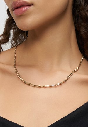 Close-up van de hals van een vrouw die een delicate gouden ketting met langwerpige schakels draagt, gecombineerd met een zwarte top.