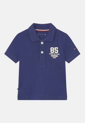 Polo bleu marine en coton texturé avec un col, trois boutons, et le numéro "85" ainsi que le nom de la marque imprimés en blanc sur le devant.