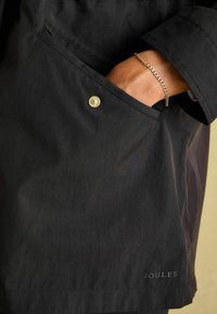 Czarna wodoodporna kurtka z przednią kieszenią z złotym zapięciem na napę, teksturowanym materiałem i subtelnym logo marki blisko krawędzi.