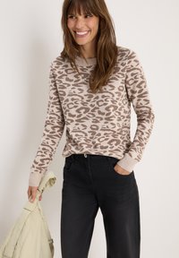 Pull beige avec imprimé léopard brun, col rond et poignets côtelés, associé à un pantalon noir. Le mannequin tient un sac de couleur claire.
