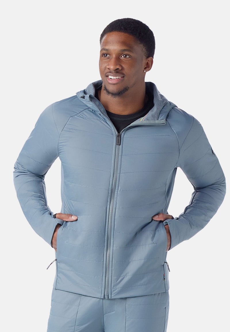 Ljusgrå pufferjacka med huva, med quiltad textur, dragkedjestängning och sidofickor. Materialet verkar slätt och hållbart.