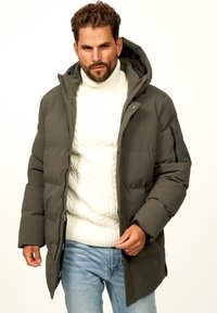 Dunkelgrüne Steppjacke mit Kapuze, verfügt über einen Reißverschluss und seitliche Taschen, getragen über einem weißen Zopfmustersweater und hellblauen Jeans.