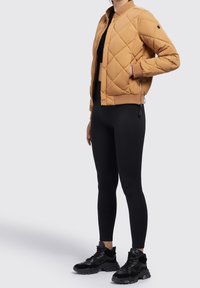 Personne portant une veste matelassée beige, un crop top noir, des leggings noirs et des baskets épaisses noires, debout avec une main dans la poche.