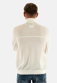 Maglione chiaro a maglia con colletto alto, caratterizzato da un dettaglio di cucitura orizzontale e una piccola etichetta sul retro. Indossato sopra jeans blu.