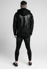 Giacca nera in pelle con cappuccio, dal design aderente e finitura testurizzata, abbinata a jeans skinny neri e sneakers nere.