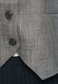 Γκρι σακάκι από ύφασμα τύπου ψαροκόκαλο με μαύρα κουμπιά, που διαθέτει κλασικό πέτο με εγκοπή και διακριτική υφή, φορεμένο πάνω από μαύρο πλισέ ρούχο.