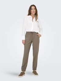 Chemise blanche à manches longues associée à un pantalon de jogging beige ample avec une taille élastique. Des sneakers olive complètent la tenue.