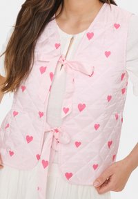 Padded brezrokavnik v svetlo roza barvi z темно roza srčnimi vzorci. Ima vezanje spredaj in gladko, sijočo teksturo.