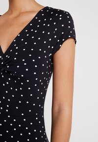 Body noir à pois blancs, col en V, manches courtes et détail torsadé à l'avant, en tissu extensible.