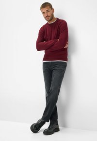 Bordeauxfarbener Pullover mit einem weißen Kragen, kombiniert mit dunkelgrauen Jeans und schwarzen Schuhen. Das Model steht mit verschränkten Armen vor einem schlichten Hintergrund.
