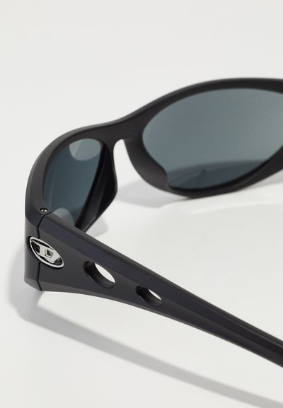 Lunettes de soleil noires avec des verres sombres et une finition mate, présentant un design découpé sur les tempes et un accent métallique sur le côté.