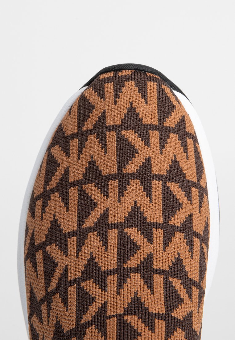 Zapatilla de punto marrón y naranja con un patrón geométrico de flechas, suela blanca y parte superior texturizada. Presenta una punta redondeada y acentos en contraste.