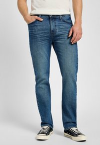 Jeans de mezclilla azul con corte recto, desgaste ligero, diseño de cinco bolsillos y cierre de cremallera, combinados con zapatillas negras.