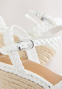 Gros plan de sandales compensées en cuir tressé blanc avec boucles argentées et semelles en jute tissé.