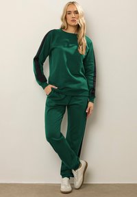 Felpa verde e pantaloni abbinati con dettagli neri. Tessuto liscio, scollatura a girocollo e vestibilità comoda. Le scarpe sono bianche con suola in gomma.