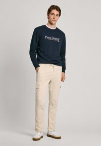 Námornícka mikina s bielym logom "Pepe Jeans", v kombinácii s svetlo béžovými cargo nohavicami s vreckami, nosená s bielymi teniskami a hnedými podrážkami.