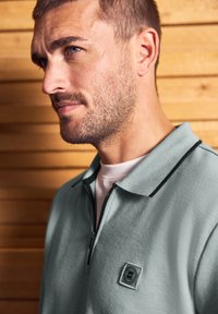 Muž s krátkými vlasy a vousy, oblečený v světle šedém polo triku na zip s logem, stojící proti dřevěné latové zdi.