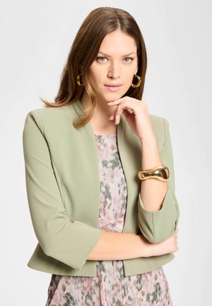 Vrouw met lichtbruin haar draagt een saliegroene cropped blazer over een bloemenjurk, gecombineerd met gouden oorringen en een chunky gouden armband.