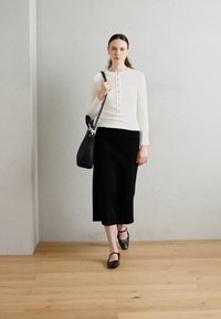 Biela blúzka s dlhým rukávom a detailmi na gombíky, skombinovaná s čiernou sukňou po kolená. Čierna kabelka a topánky na remienky dopĺňajú outfit.
