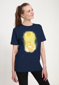Maglietta blu navy con un sole in stile cartone animato e una creatura gialla e pelosa con un volto sorridente, su uno sfondo di alone giallo chiaro.