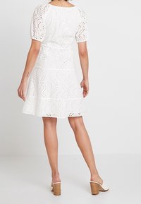 Femme portant une robe en dentelle blanche à manches bouffantes et des mules à talons blancs, se tenant devant un fond clair uni, de dos face au spectateur.