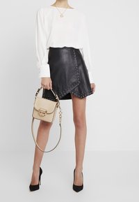 Jupe portefeuille en cuir noir avec des accents cloutés, associée à un chemisier blanc. Tenant un sac à main en crème texturé. Chaussures à talons hauts noires.